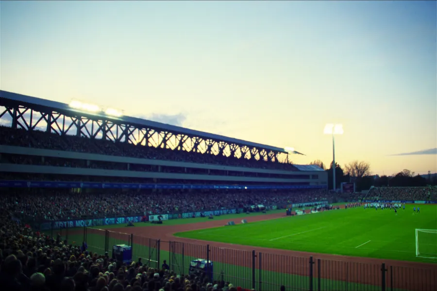 A floodlit stadium at dusk with a brightly lit pitch and a full crowd